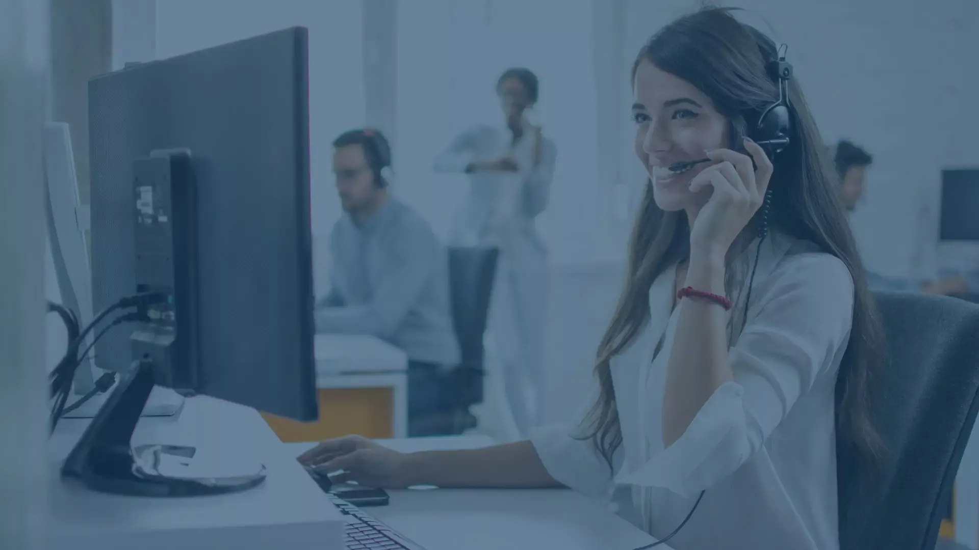 Lachende IT Service Desk Mitarbeiterin mit Headset am Computer bei der Arbeit. Fokus auf Kundensupport, Erreichbarkeit und Performance im modernen Service Center. Im Hintergrund sind weitere Kollegen sichtbar.