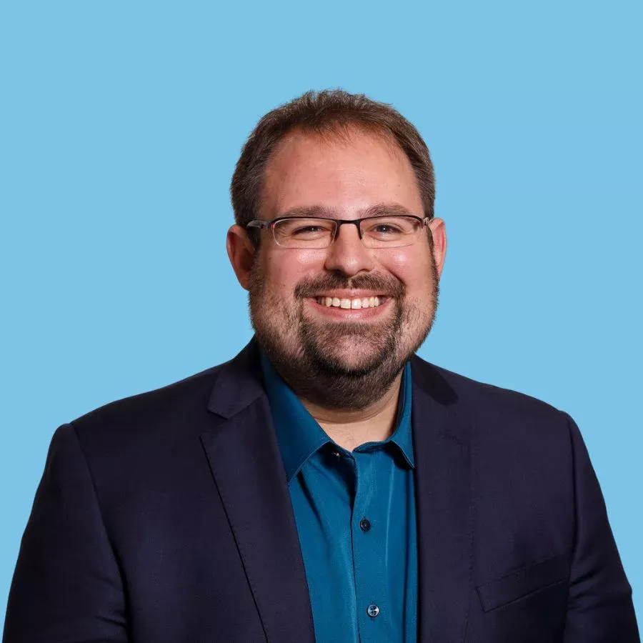 Porträtfoto eines lächelnden Mannes (Jerome Drees) im Business-Outfit vor hellblauem Hintergrund.